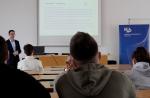 Mężczyzna prowadzi szkolenie dla studentów. Studenci siedzą na sali wykładowej przy biurkach. Na ekranie wyświetlana jest prezentacja, po prawej stronie stoi niebieski baner Krajowej Administracji Skarbowej.